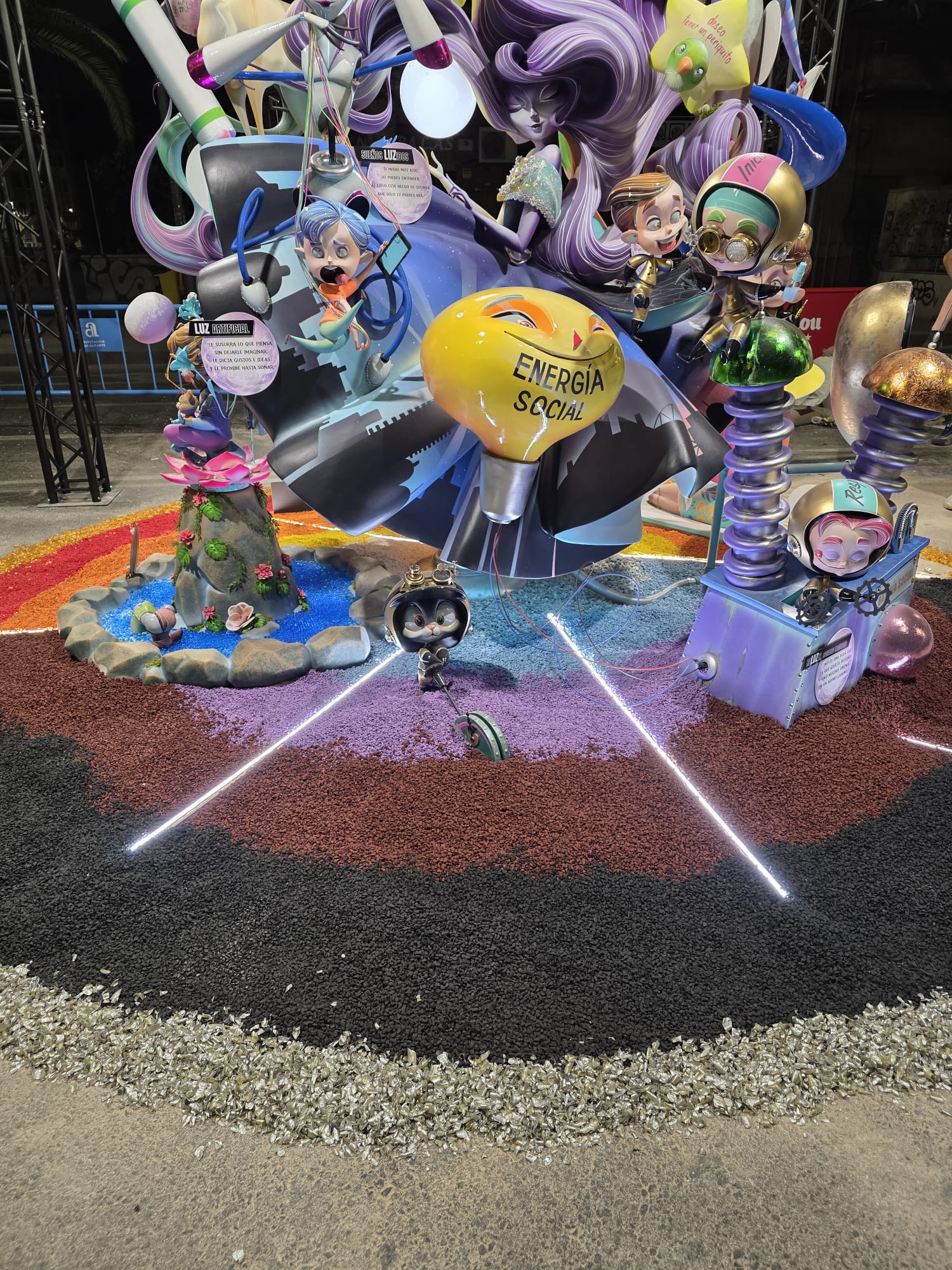 Escena de la Hoguera Infantil Diputació-Renfe 2026 donde la iluminación LED en tonos fríos realza la composición y aporta profundidad al conjunto. La decoración combina una gama cromática de azules, lilas, violetas y negros, creando un ambiente tecnológico y futurista que refuerza el mensaje artístico del monumento. El suelo se ha trabajado con áridos decorativos de diferentes colores, mientras que las líneas de luz LED delimitan la escena y dirigen la mirada hacia el elemento central, logrando un efecto visual moderno y lleno de contraste.